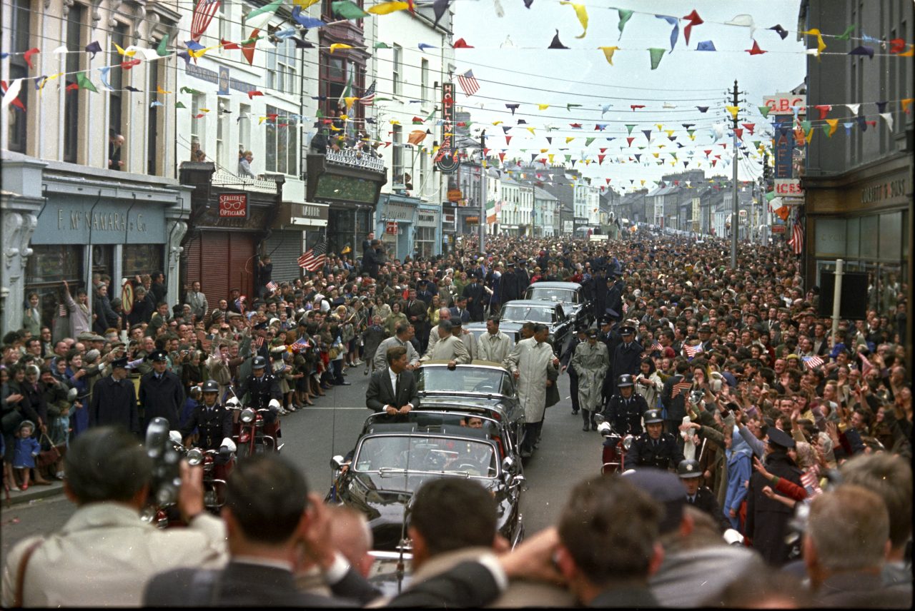 Colour photo of JFK motorcade travelling through Williamsgate Street, Galway in 1963 with crowds of people on either side of the street cheering him on. 